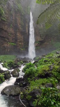 Kapas Biru Waterfall surrounded by lush tropical rainforest in East Java, Indonesia. Scenic jungle landscape with cascading water, mist, and vibrant green vegetation.