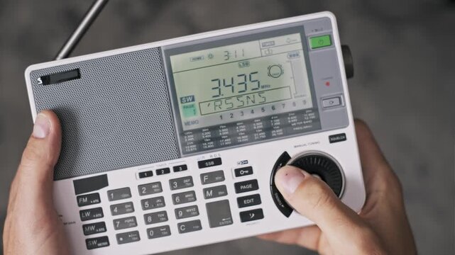Close-up of hands tuning a modern portable shortwave radio with a manual dial. The shot reveals a changing digital frequency display and numeric keypad against a grey background.