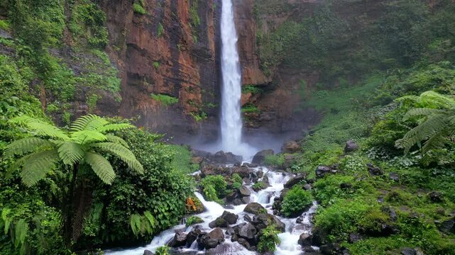 Aerial view of Kapas Biru Waterfall surrounded by lush tropical rainforest in East Java, Indonesia. Scenic jungle landscape with cascading water, mist, and vibrant green vegetation.