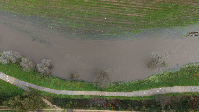 Aerial view of urban river flooding with eco trail next to it, showing muddy water overflowing its banks into surrounding green fields and vegetation in Portugal.