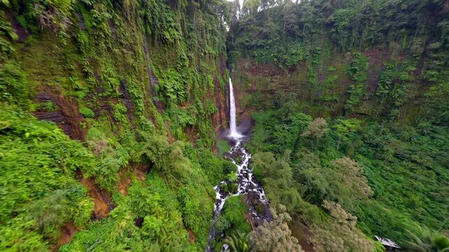 Cinematic FPV drone flight toward Kapas Biru Waterfall through lush tropical rainforest in East Java, Indonesia. Dramatic jungle landscape with a tall cascading waterfall and mist.