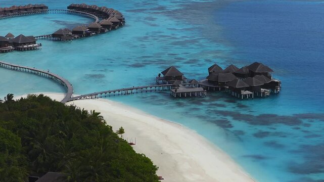 Aerial view of a Maldives resort island, S shaped and circular jetties link overwater bungalows, white sand beach and palms, reef patches, people walk in bright daylight