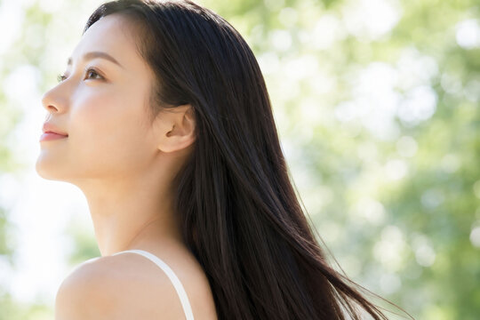 新緑のやわらかな自然光を浴びながら横顔で上を見つめるロングヘアの若い女性の透明感ある素肌と美しい髪を表現した爽やかな美容ポートレート