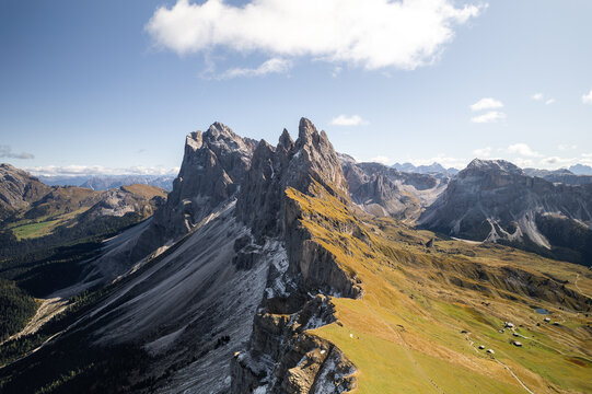 Aerial view of the dramatic Seceda mountain ridge with its jagged rocky peaks and lush green alpine slopes under a clear blue sky in Urtijei, Trentino-Alto Adige, Italy.