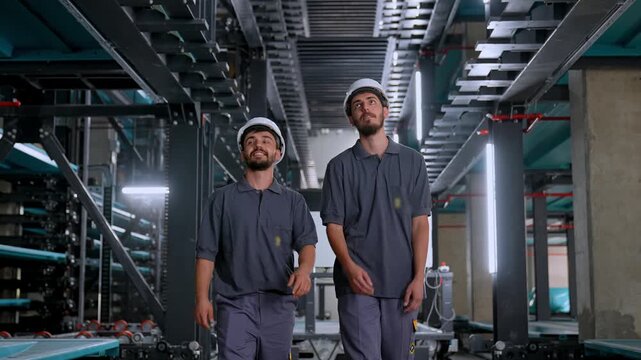 Two helmeted engineers inspect an automated parking system, looking up and pointing at overhead mechanisms while coordinating technical work inside a modern parking facility.