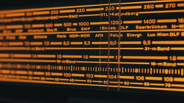 Macro pan across a vintage radio tuner analog display with a warm orange glow. The scale shows wavebands, MHz numbers, and broadcast station names on a dark background.