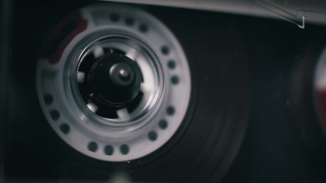 Close-up of a transparent audio cassette playing in a player. White spools rotate steadily, winding magnetic film in a dark setting, evoking 90s mixtape nostalgia.