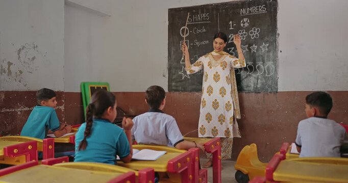 Indian young adult female talk speak teach to girl boy group class room board indoor rural area teen child wear uniform sitting on bench look tutor follow learn poem action moves enjoy day desi life