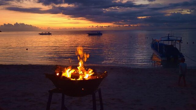 A serene Bali sunset captured around a glowing firepit on the beach. Traditional wooden boats sit anchored just offshore, silhouetted against a sky ablaze with amber and rose.