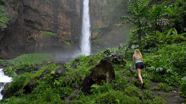 Young woman hiking through lush tropical jungle toward Kapas Biru Waterfall in East Java, Indonesia. Mist rises from the powerful cascade beneath towering cliffs.