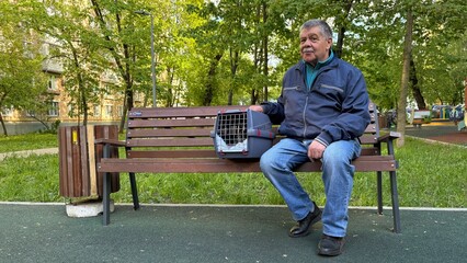 Fototapeta premium Retired man with his pet in transport cage on a walk. Happy senior man sitting on bench with cat carrier box.