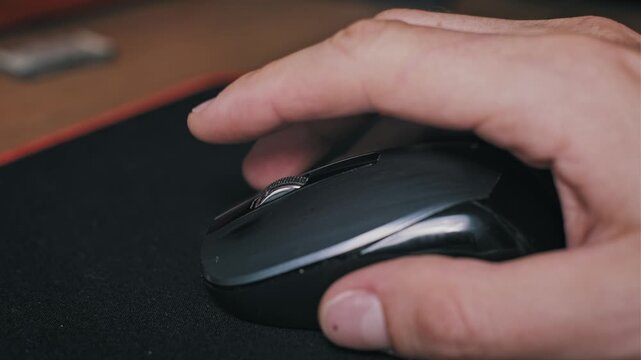Side view of a male hand scrolling the metal wheel of a black computer mouse on a wooden table. The scene represents office work, online browsing, and efficient document navigation.