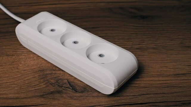 Side angle view of a male hand unplugging a white charger adapter from a white three-way extension block. The action on a dark wood table disconnects a device to conserve energy.