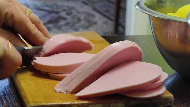 Side view of female hands with red nails using a large knife to slice pink bologna sausage on a wooden board. This home kitchen scene depicts ingredient preparation for a family meal.