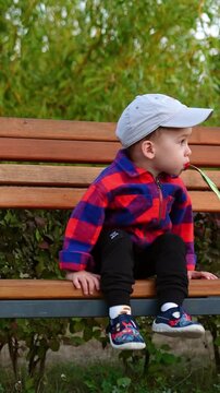Beautiful kid in cap and checkered shirt gets off the bench. Toddler drinks from doy pack in the park. Vertical video
