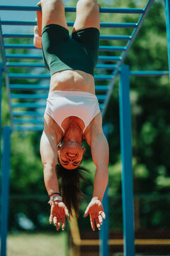 Athletic woman hangs upside down from blue monkey bars smiling and reaching toward the ground. Outdoor fitness scene shows strength and playful exercise on a sunny day.