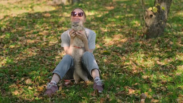 Woman holds cat gently while sitting under trees shade. She hugs her pet tenderly. Cat looks calm, eyes half-closed in trust. Sunlight filters through leaves, soft and warm.