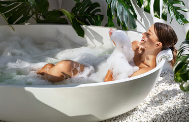 Naklejka premium A smiling young Caucasian woman enjoys a serene wellness vibe in a bathtub filled with bubbles and bright natural light