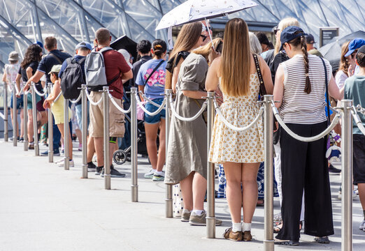People stand in line &mdash; alone, as a couple, in a group, with family or friends &mdash; between stainless steel queue poles with grey ropes at the entrance of a tourist site, on a sunny summer day.