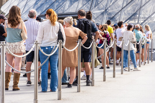 People stand in line &mdash; alone, as a couple, in a group, with family or friends &mdash; between stainless steel queue poles with grey ropes at the entrance of a tourist site, on a sunny summer day.