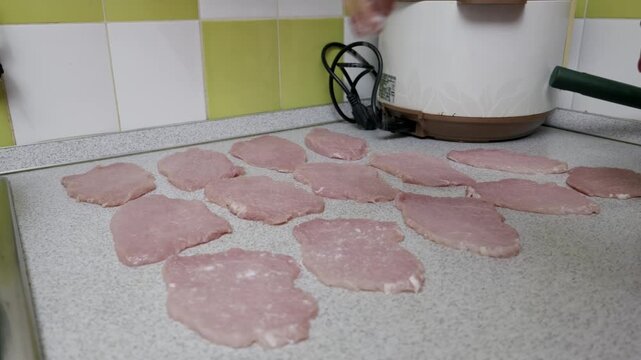 Female hand sprinkles salt over thin slices of raw pork laid out on a kitchen countertop. This close-up shot captures a moment of home food preparation with a tiled background.