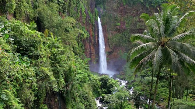 Aerial view of Kapas Biru Waterfall surrounded by lush tropical rainforest in East Java, Indonesia. Scenic jungle landscape with cascading water, mist, and vibrant green vegetation.