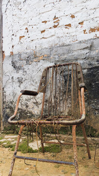 Old rusty metal chair with broken woven strings in front of weathered cracked white wall with peeling paint. Abandoned vintage furniture with corrosion, decay, and rustic texture perfect for grunge