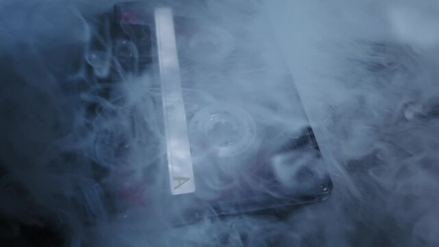 Audio cassette tape with a white label lies on a dark, reflective surface. Mysterious white smoke billows and swirls around the retro object, creating a moody atmosphere.