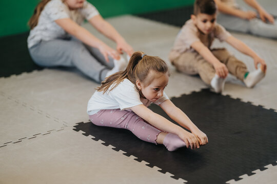 Young kids sitting on padded mats stretching forward to touch their toes in a group exercise. A small class of children practices seated stretches together on interlocking floor mats.