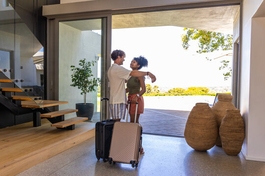 Couple standing near sliding glass door, embracing with wheeled suitcases on terrazzo floor