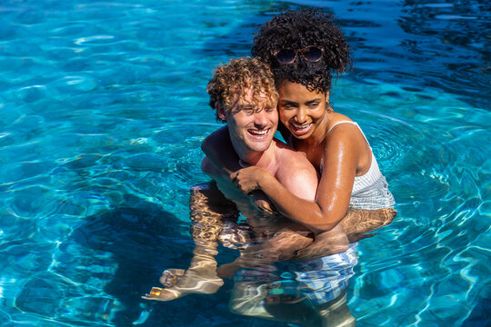 Diverse couple smiling, embracing in outdoor pool with sunglasses, swimwear and rippling water