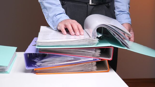 Person's hands organizing a stack of colorful ring binders and file folders filled with important paperwork, managing business documents and office administration tasks on a white desk