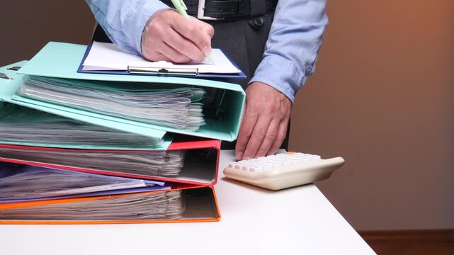 Person's hands busy calculating data on a calculator and writing notes on a clipboard, while surrounded by stack of folders filled with important business and financial documents on a desk