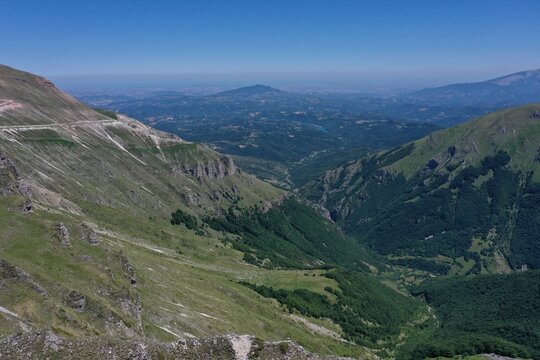 Aerial view of the rugged Sibillini Mountains with steep green slopes, rocky ridges, and deep valleys under a clear blue sky in Montemonaco, Marche, Italy.