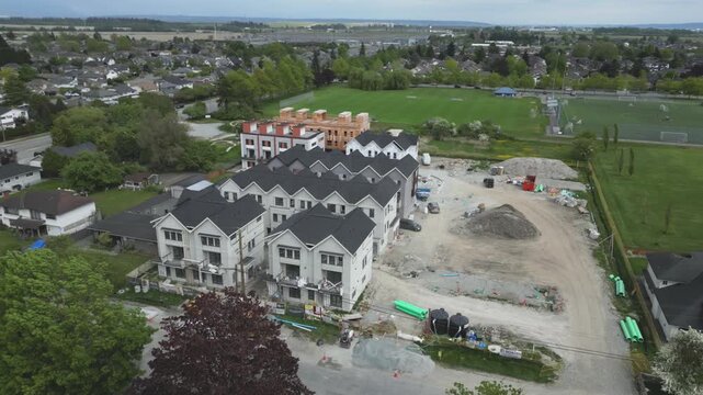 High-angle aerial perspective orbits right around sprawling residential development site showcasing newly built townhouses ranging from raw wooden framing to completed white siding black shingled roof