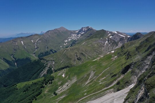 Aerial view of the rugged Sibillini Mountains with green slopes, patches of snow, and rocky peaks under a clear blue sky in Montemonaco, Marche, Italy.