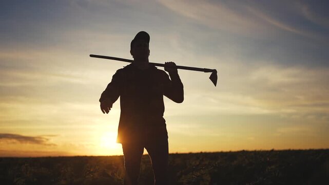 A man stands in a field holding a hoe against a sunset backdrop. The silhouette of the man and the hoe contrasts with the colorful sky and distant landscape.