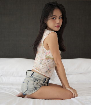 Young Asian woman wearing a floral crop top and denim shorts kneels on white bedding while looking over her shoulder