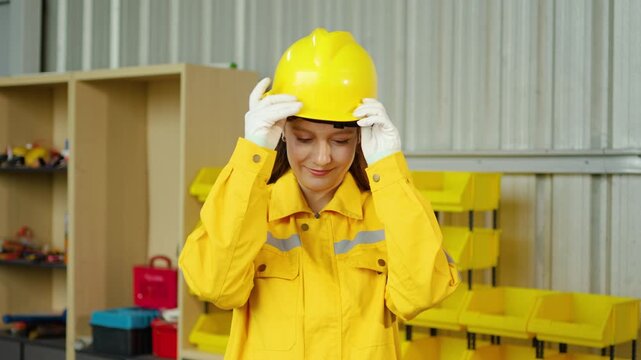 Confident female industrial engineer in a yellow uniform taking off her safety hard hat and smiling inside a manufacturing warehouse facility, representing professional women in the labor workforce.