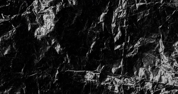 Close-up of shiny dark crinkled surface with sharp folds and reflective highlights. Smooth continuous rotation reveals dynamic shifts in deep shadows and bright specular reflections.