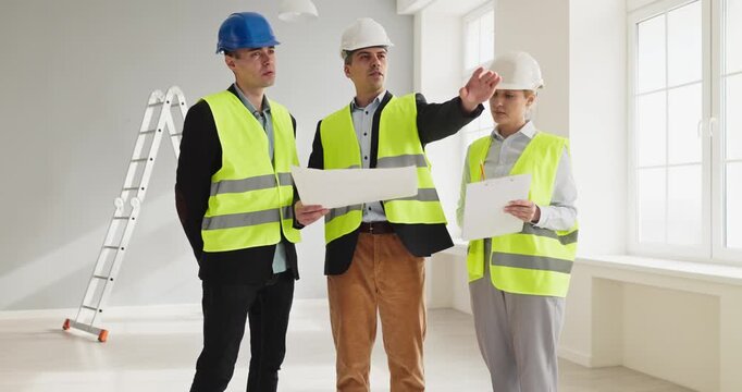 Workers engineers and architect discuss planning. Wearing helmets and vests, the team leads an interior renovation inspection and discusses quality. Teamwork in renovation with blueprints.