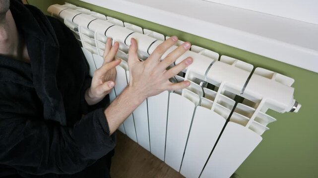 A frozen man in a warm bathrobe touches a white wall radiator to check the temperature and hugs himself. Concept of heating season problems, rising energy prices, and cold indoor climate.