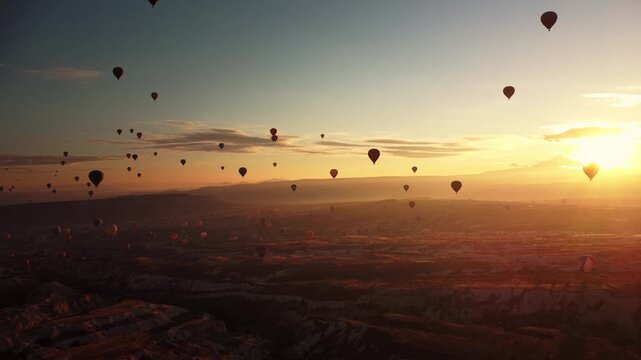 Multitude of balloons drifting over open land during dramatic sunset scene filmed by drone. Aerial view of dozens aerostats casting long shadows across rocky fields and valleys with sun on horizon.