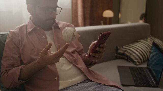 Slowmo shot of Indian young man sitting on sofa, using smartphone and tossing baseball while working from home. Ideal for videos about remote work procrastination or sports betting apps