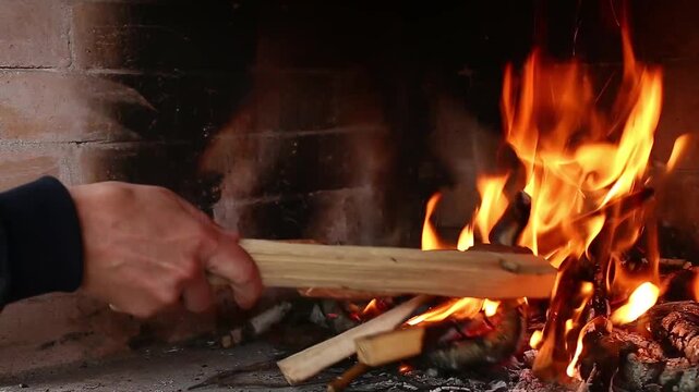 Man's hand adding firewood to a burning brick fireplace from the left side, close-up front view, dark interior background