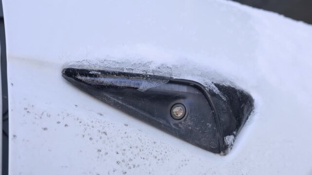A white electric vehicle side repeater camera sits covered in frozen ice and snow during a cold winter day. Concept of winter driving challenges and electric car performance in cold weather.