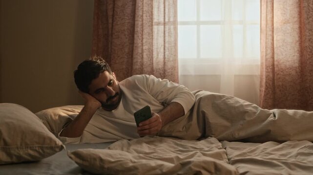 Medium long shot of young Indian man lying in bed, scrolling on smartphone after waking up. Ideal for mobile application promotions, digital wellness campaigns