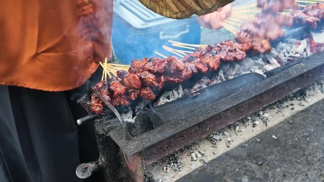 Grilled meat sizzles over smoldering coals. Vendor shields food with a bamboo umbrella. Smoke curls around the skewers. Hands adjust sticks over open flame. Street food scene, casual, warm, hungry