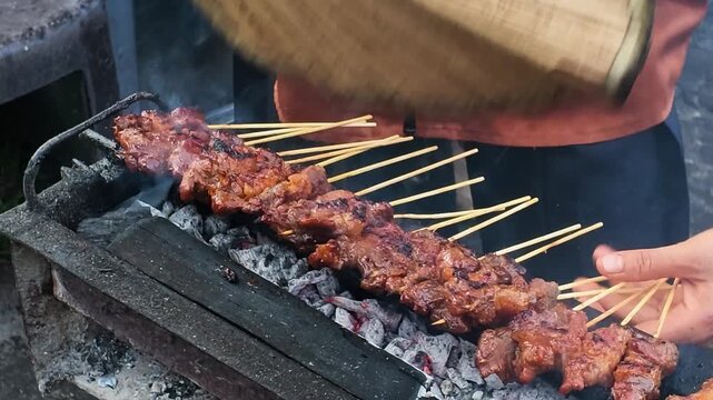 Satay skewers sizzle over glowing embers barbecue the traditional way using hand fan in Malaysia. Hands adjust skewers, checking doneness. Smoke curls upward, filling the air. Perfect for food, street