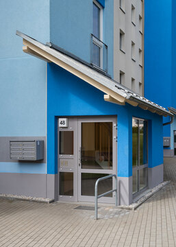 Modern residential apartment building entrance featuring vibrant blue and grey plastered walls, a glass door, mailboxes, and a striking angled canopy roof.
